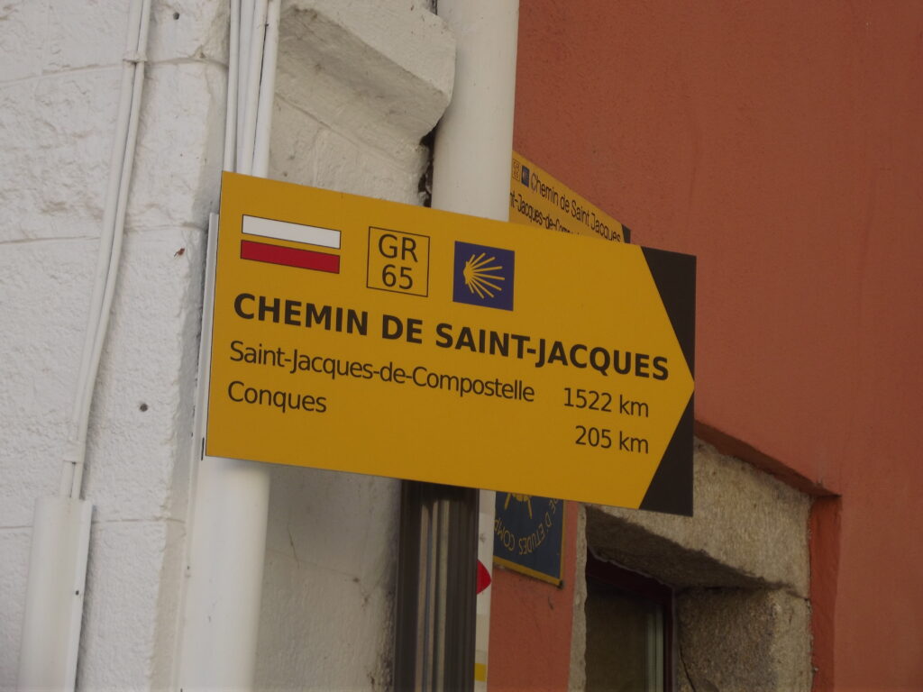 Panneau de randonnée indiquant les données suivantes:
CHEMIN DE SAINT-JACQUES
Saint-Jacques-de-Compostelle 1522 km
Conques 205 km